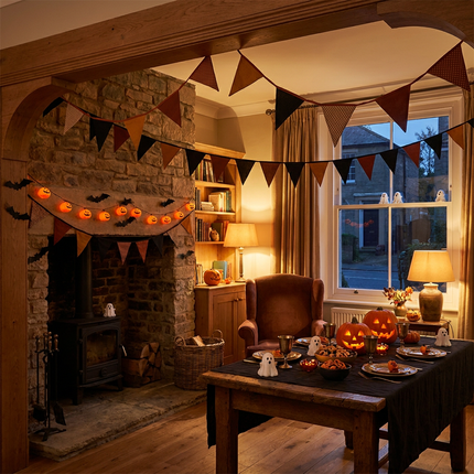 Cozy Dutch living room dressed with Halloween slingers, bat bunting and pumpkin garlands above a party table lit by warm evening light.